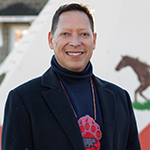 John Sylliboy, souriant, devant un tipi blanc décoré d'un motif rouge et de l'image d'un cheval.