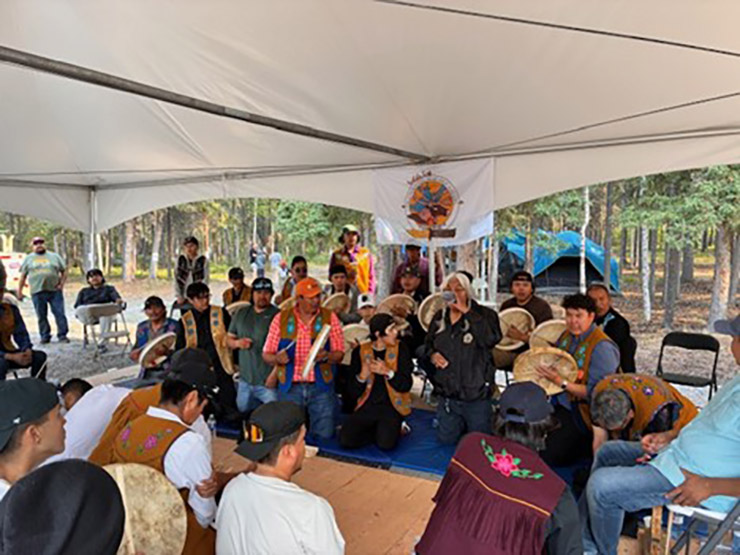 This picture shows members of the Yellowknives Dene First Nation kneeling in 2 groups, facing each other, under a tent awning. Many of the people are drumming.
