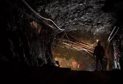 Photograph of the silhouette of a person in personal protective equipment in an underground mine tunnel. The tunnel is hollowed out rock with wires and pipes along the top.