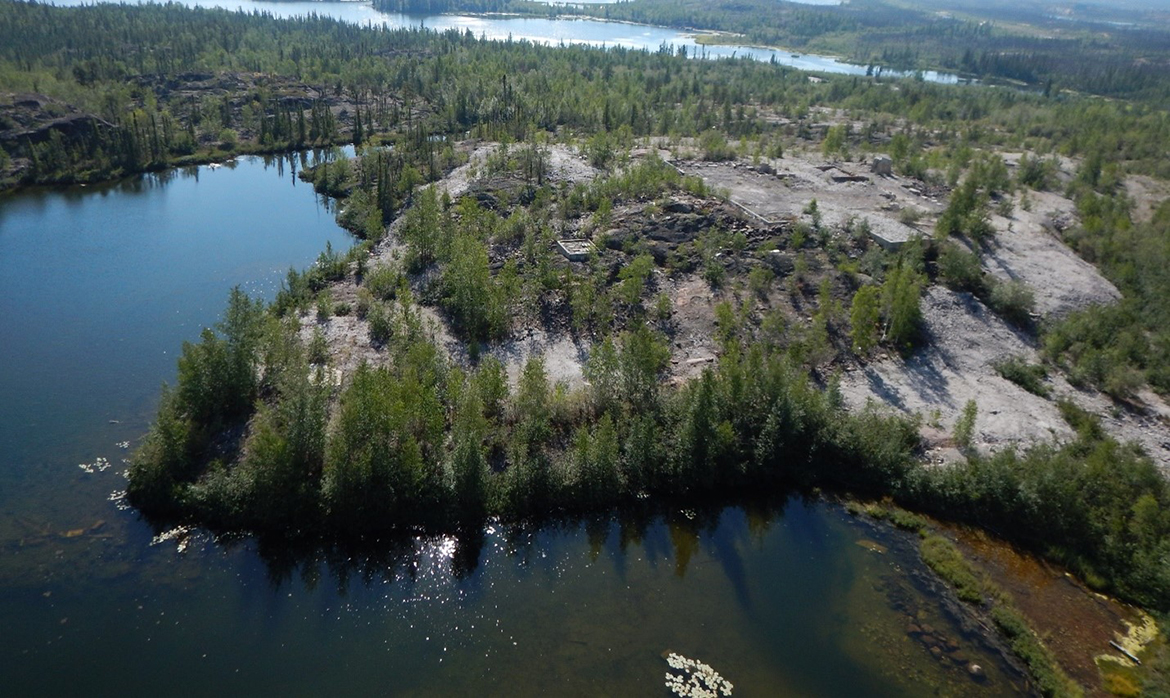 An aerial view of the Rayrock Mine site. The photo shows a rocky terrain with pine trees, a lakeshore visible in the foreground and the background. A large section of land where the mine site once stood is cleared of trees, leaving the rock and a few buildings visible.