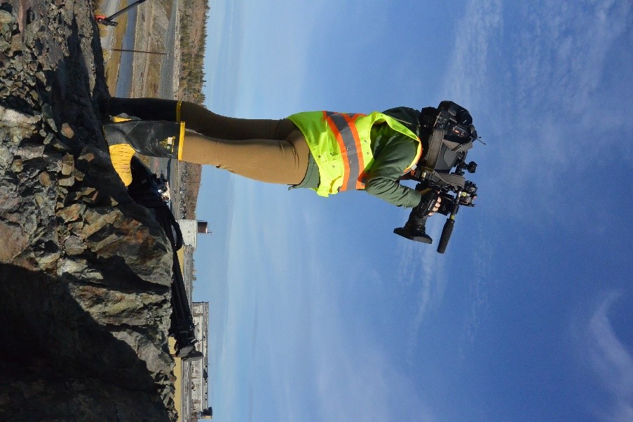 A reporter dressed in personal protective equipment and holding a video stands on a rocky outcrop. Dilapidated buildings and a creek are visible in the background.