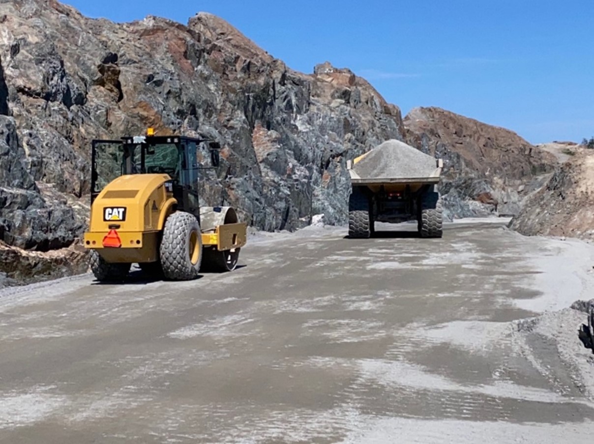 Heavy equipment (a roller and a dump truck carrying aggregate material) drive on an unpaved road the lower portion of the AR1 Pad in front a rocky outcrop